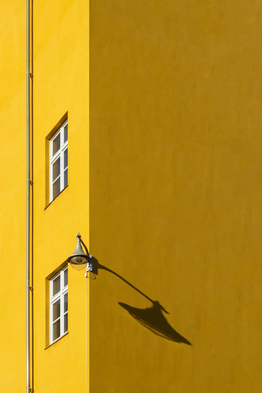 A sunlit ochre facade with white-framed windows and a street lamp casting a long shadow